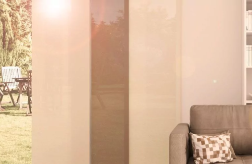 Cremefarbiger Flächenvorhang an einem hohen Fenster hinter einem hellbraunen Sofa